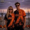 Collection shot of horror Hawaiian shirts in all available colors, featuring haunted house, bats, spider webs, and jack-o'-lantern print, worn by a couple against a dynamic background