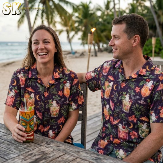 A couple wearing dark purple halloween hawaiian shirts printed with ghost-filled tropical cocktails, hibiscus flowers, palm leaves, and bats in a watercolor style.