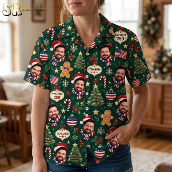 A close-up view of a woman wearing the dark green shirt, bringing out the finer details of the cartoonish motifs including the smiling Santa-hat face, holly sprigs with red berries, gold stars, and the AMERICA 250 text on the circular ornaments.