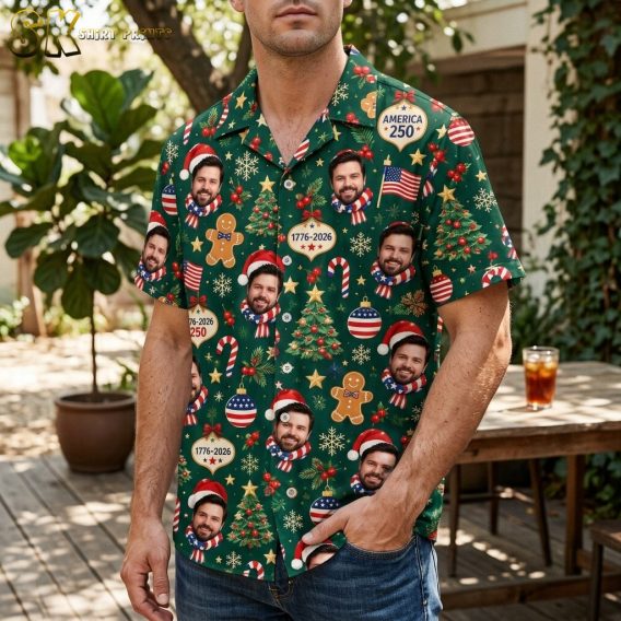 A man wearing the dark green short-sleeve button-up shirt, showing how the patriotic Christmas pattern looks when worn, with the smiling Santa-hat faces, gingerbread men, and AMERICA 250 ornaments visible across the chest and body.