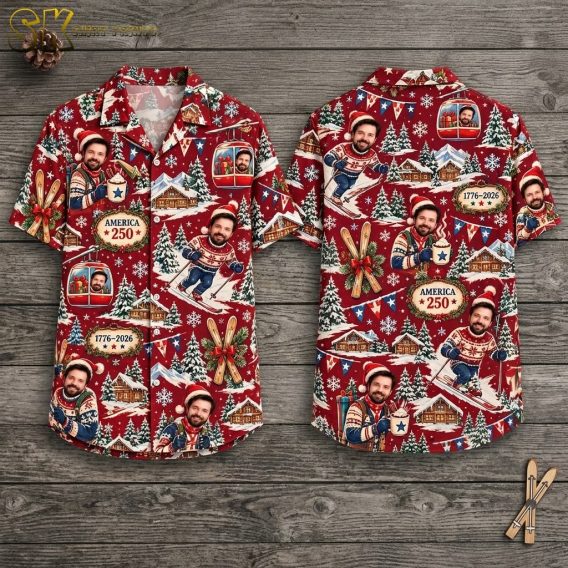Both sides of a deep-red short-sleeve button-up shirt displayed on a wooden hanger, showing repeated printed faces of a smiling man in a Santa hat skiing, gondola cabins, crossed skis with a red ribbon, and emblem plaques reading AMERICA 250 and 1776-2026.