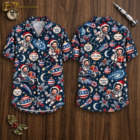 Front and back hanger view of a dark navy blue short-sleeve button-up shirt covered in an all-over pattern of astronauts in Santa hats, Christmas ornaments, candy canes, stars, planets, and America 250 badges with the dates 1776-2026.