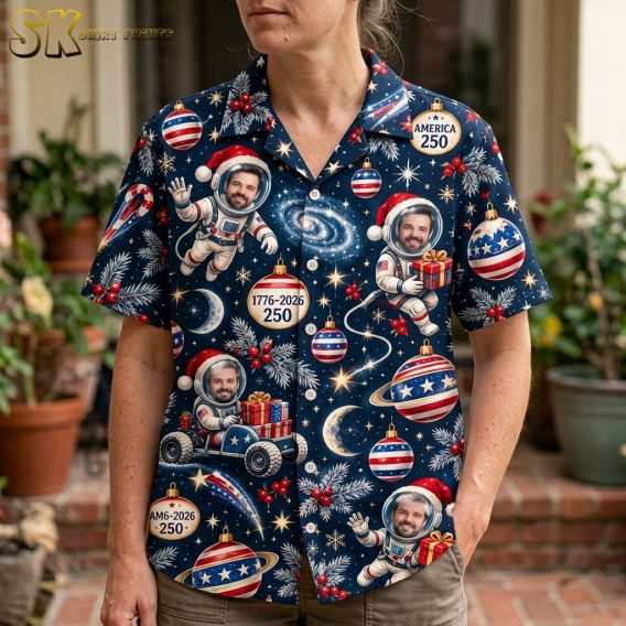 A close-up view of a woman wearing the navy shirt, bringing out the detailed cartoon artwork of astronauts with bearded faces visible inside their helmets, Santa hats, snowflakes, crescent moons, and America 250 ornament badges.