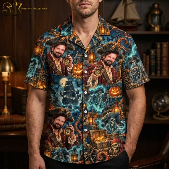 A man wearing a mens halloween hawaiian shirt stands indoors against a dark wood bookshelf backdrop, with the repeating pirate portrait and jack-o'-lantern motifs clearly visible across the chest.