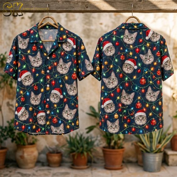 Two christmas aloha shirts hanging from a wooden rod outdoors, showing the front of one and the back of the other, with the navy cat-face and string-light all-over print clearly visible against a rustic stone wall.
