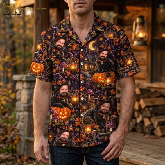 A man wearing a short-sleeve button-up shirt with an all-over Halloween print featuring a black cat with a man's face, glowing lanterns, and jack-o'-lanterns on a dark purple background.