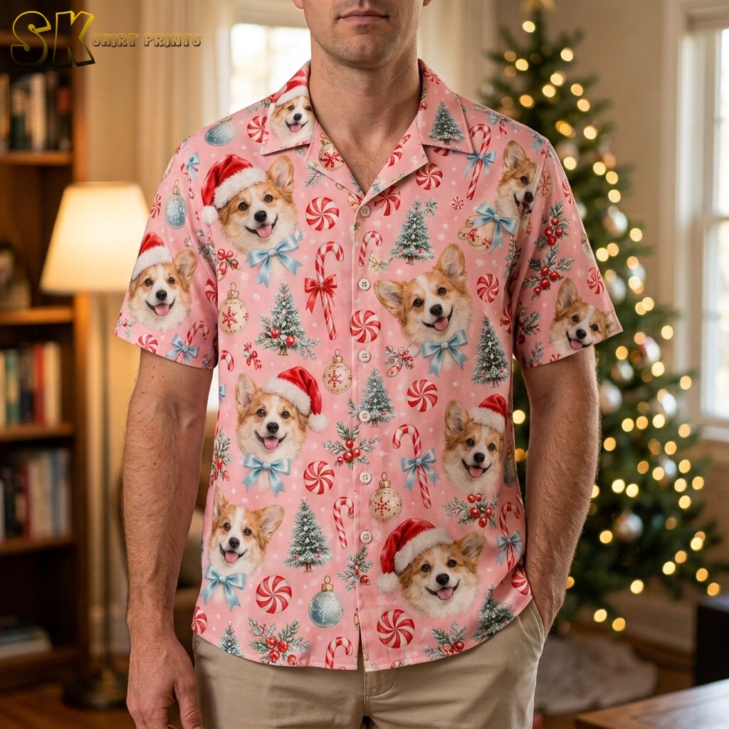 A man wearing a pink men's christmas hawaiian shirt stands in front of a lit Christmas tree, the corgi Santa hat faces and peppermint candy motifs clearly visible across the chest and sleeves.