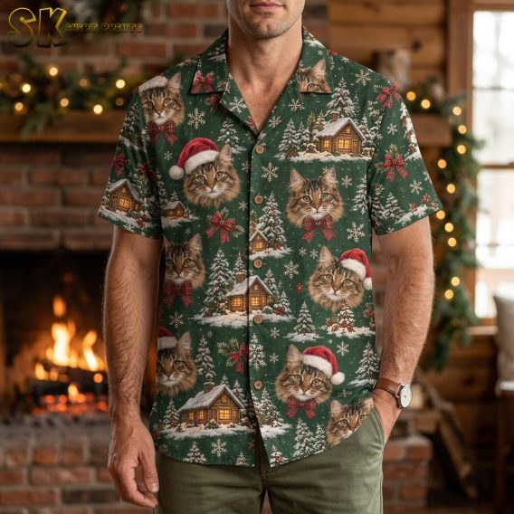 A man wearing a men's christmas hawaiian shirt stands in front of a lit stone fireplace, the green fabric covered in large tabby cat faces, some wearing Santa hats, and glowing cabin scenes between snowflakes.