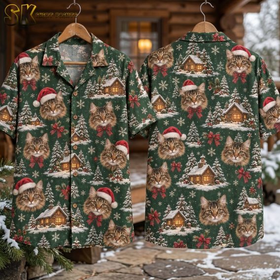 Two hangers display the front and back of a green short-sleeve button-up shirt outside a snow-dusted cabin porch, with the full repeat of tabby cat faces, red Santa hats, plaid bows, snowy pine trees, and glowing cabins visible on both sides.