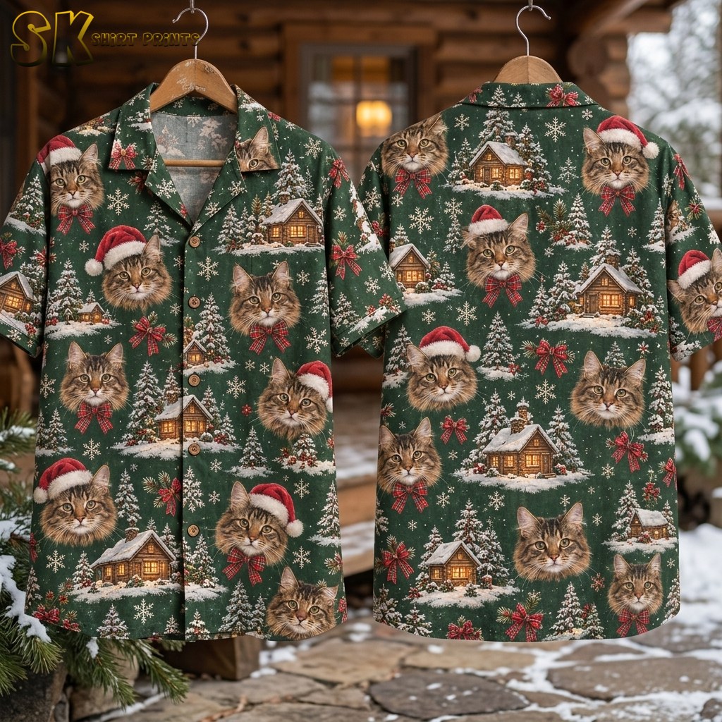 Two hangers display the front and back of a green short-sleeve button-up shirt outside a snow-dusted cabin porch, with the full repeat of tabby cat faces, red Santa hats, plaid bows, snowy pine trees, and glowing cabins visible on both sides.