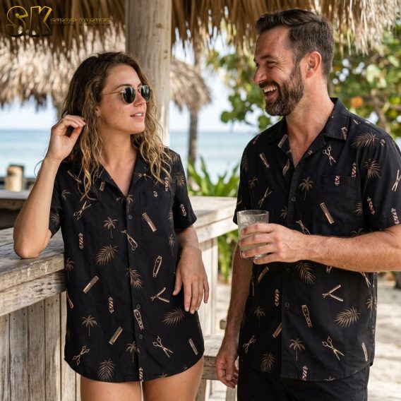 A woman and a man, both wearing the same black barber-themed shirt with palm tree and scissor prints, stand together at an open-air beachside bar.