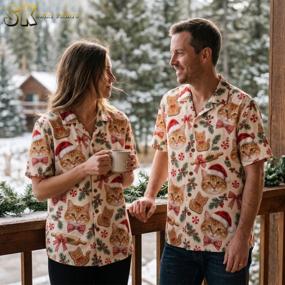 A woman and a man both wearing matching orange tabby cat Christmas Hawaiian shirts stand on a wooden porch with snow-covered pine trees behind them, smiling at each other.