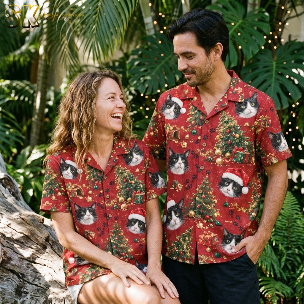 A smiling couple wearing matching red cat-print Christmas shirts stand together among tropical greenery, with tuxedo cat faces and gold ornament motifs visible on both shirts.
