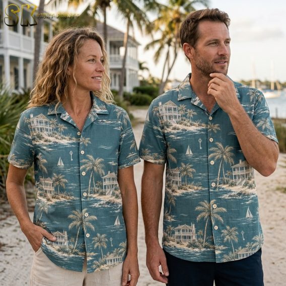 A woman and a man stand side by side on a sandy path wearing matching teal Hawaiian shirts printed with coastal houses, palm trees, and sailboats, with real palm trees and waterfront buildings visible behind them.