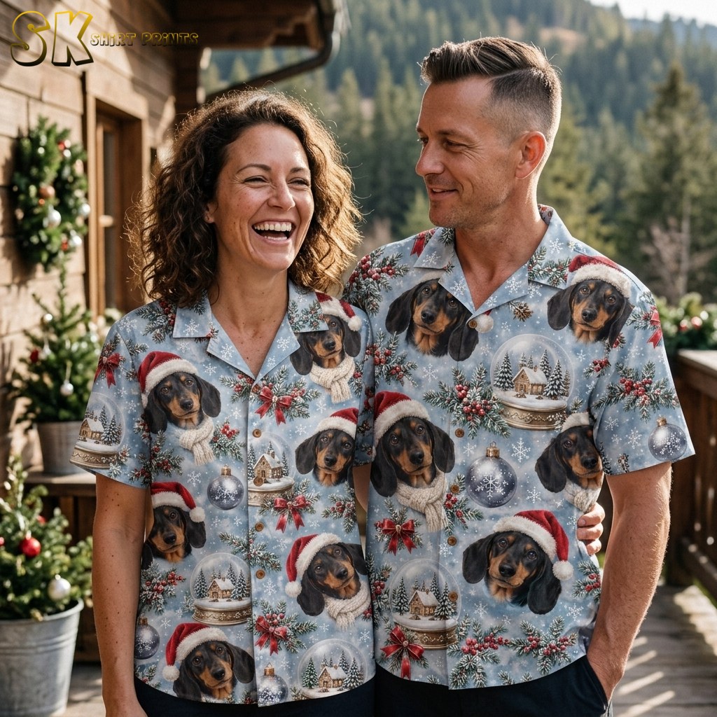 A laughing woman and a smiling man stand together on a wooden deck wearing matching dachshund Christmas shirts, with snow-dusted pine trees and holiday wreaths visible behind them.