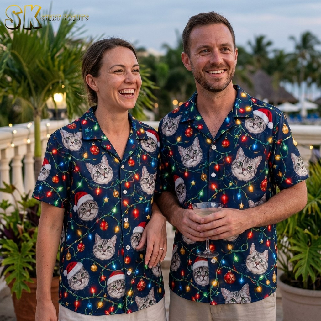 A smiling man and woman wearing matching navy hawaiian christmas shirts for family, both printed with cat faces in Santa hats and colorful string lights, standing together on an outdoor terrace with palm trees behind them.