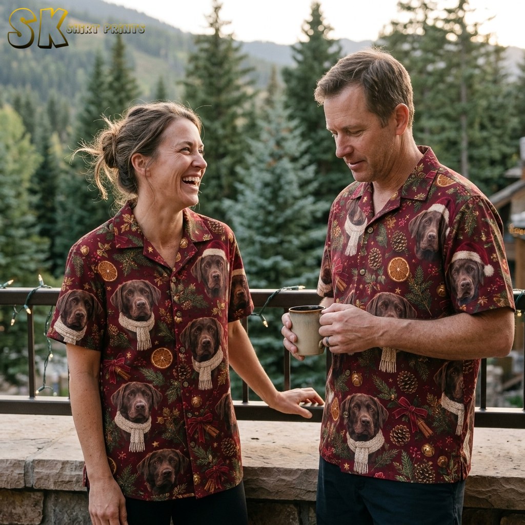A laughing woman and a smiling man both wearing the same burgundy Labrador Christmas shirt while standing on a wood-railed balcony with evergreen trees behind them.
