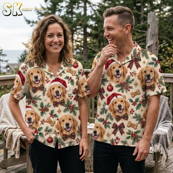 A smiling woman and man stand together on a wooden deck wearing matching Golden Retriever Christmas shirts, with pine trees and overcast sky in the background.
