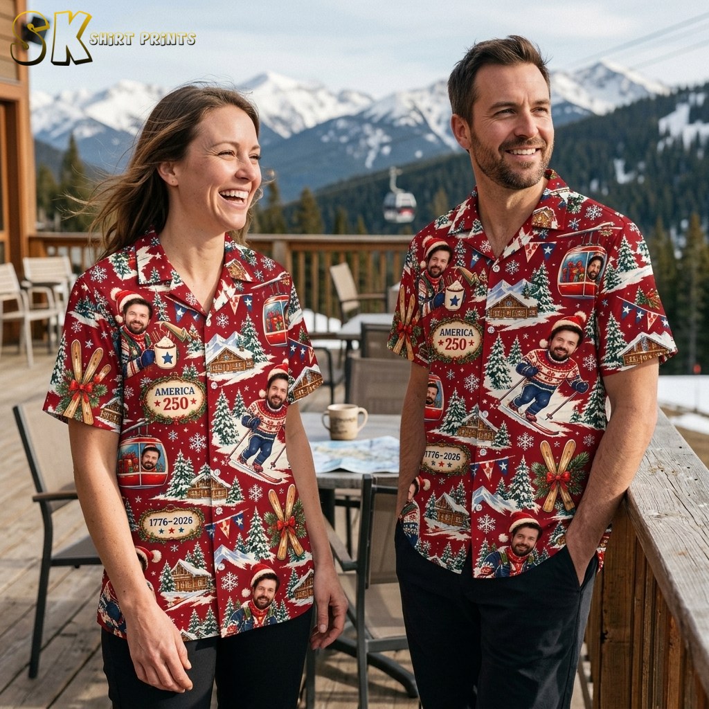 A smiling woman and man stand side by side on a snowy mountain deck, both wearing the same red winter-ski print shirt with repeated face graphics and the 1776-2026 anniversary plaque.