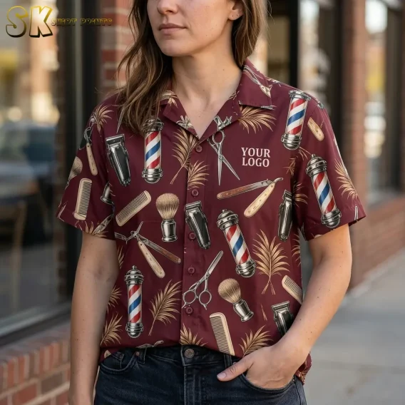 A woman wearing the maroon barber shop shirt stands on a sidewalk outside a brick building, with the 'YOUR LOGO' chest print and individual motifs including scissors, shaving brushes, and barber poles clearly visible up close.