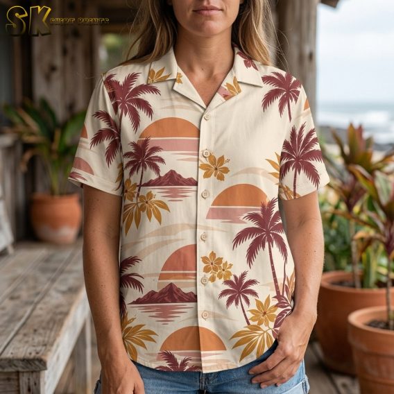 A woman stands on a weathered wooden porch near the ocean wearing the cream Hawaiian shirt, with the repeating mountain-and-sunset scenes and golden hibiscus blooms prominent across the front panels.