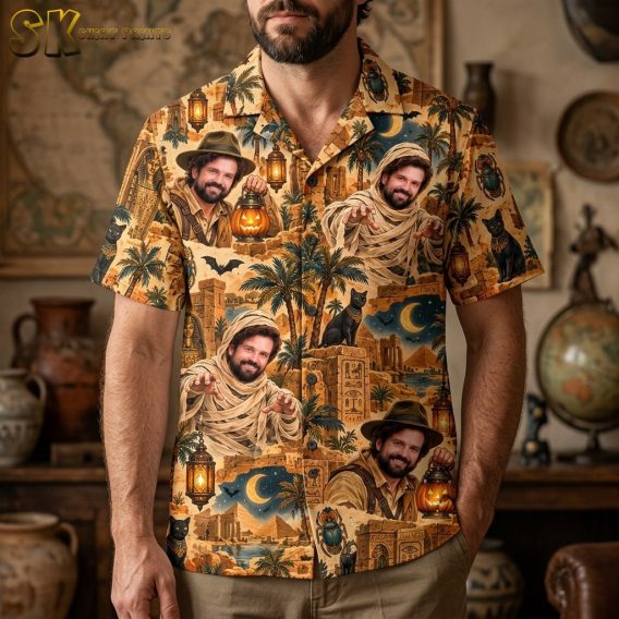 A man wearing a sandy beige short-sleeve button-up shirt printed with explorer and mummy portraits, pyramids, scarab beetles, and crescent moon motifs against a night sky.