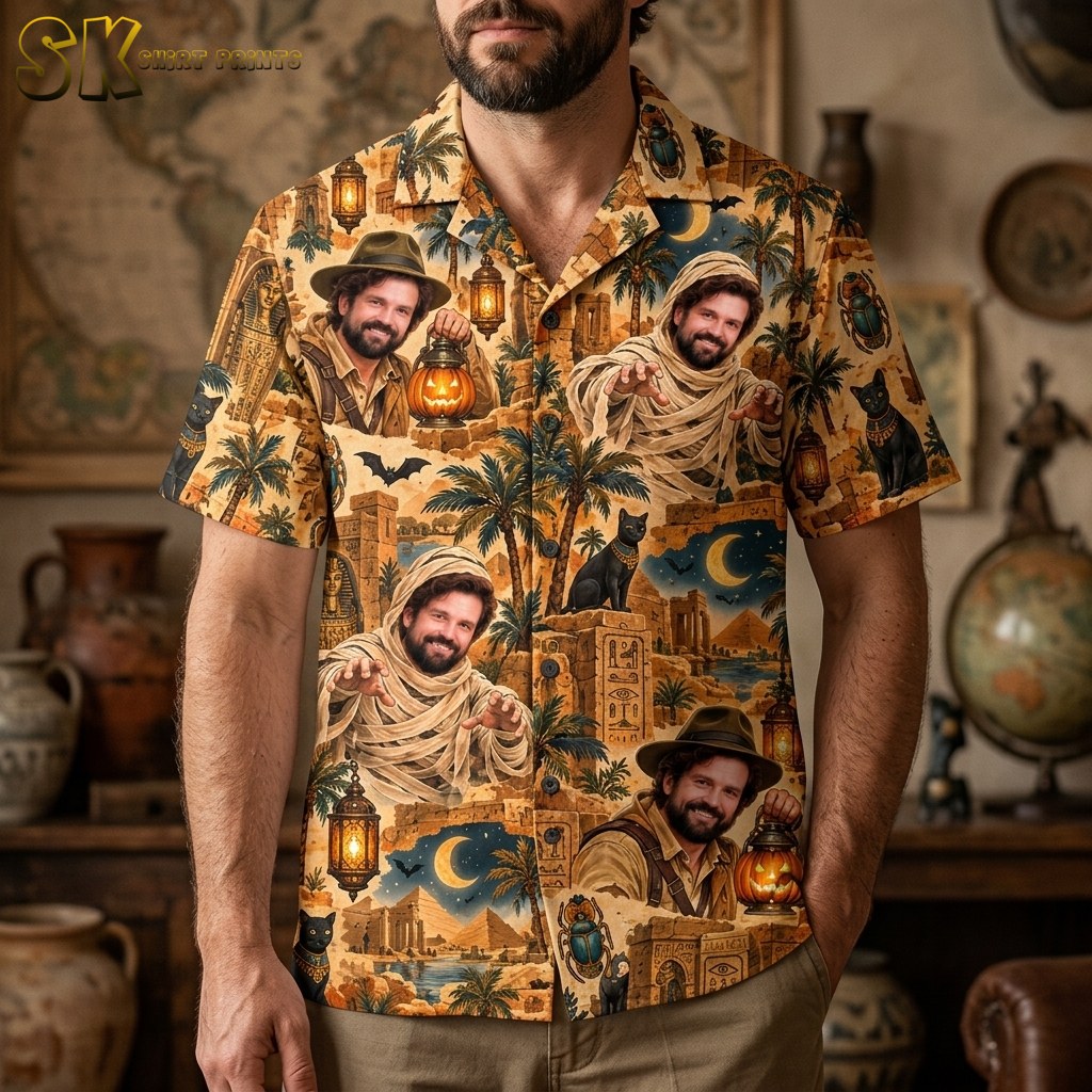 A man wearing a sandy beige short-sleeve button-up shirt printed with explorer and mummy portraits, pyramids, scarab beetles, and crescent moon motifs against a night sky.