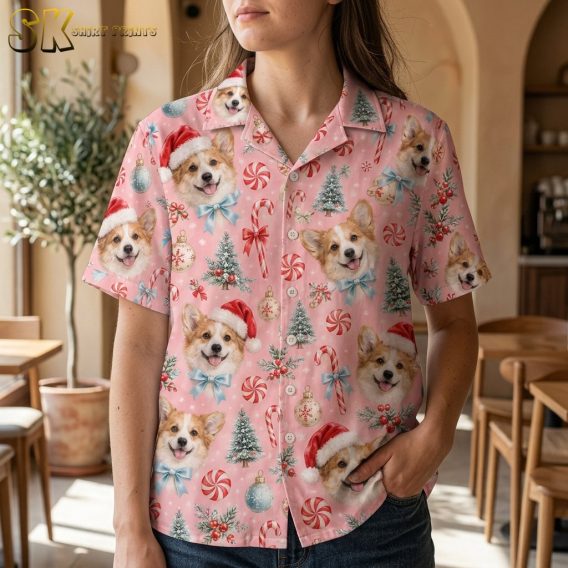A woman wearing the pink corgi-print shirt stands in a bright café interior, the realistic dog faces wearing Santa hats and blue bows filling the fabric from collar to hem.