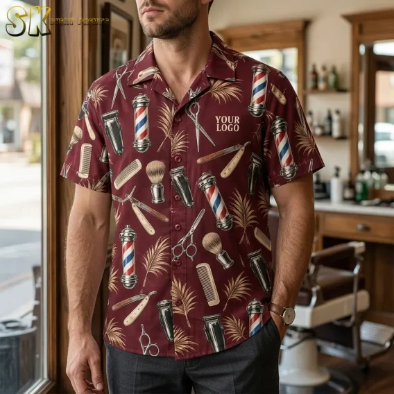 A man wearing a maroon logo hawaiian shirt printed with barber poles, straight razors, and hair clippers stands inside a traditional barbershop with wooden fixtures visible behind him.