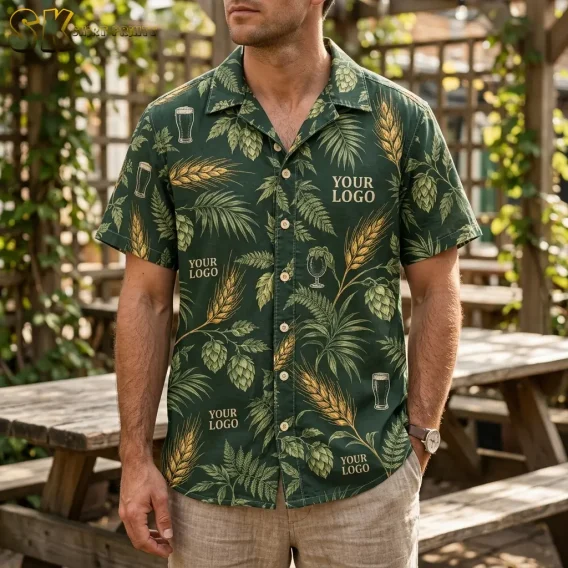 A man stands in an open-air beer garden wearing a dark green logo Hawaiian shirt printed with hop cones, barley, and beer glass motifs, with wooden picnic tables visible behind him.