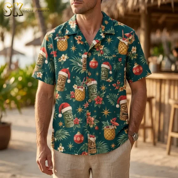 A man stands at an outdoor tiki bar wearing a mens christmas hawaiian shirt, the teal fabric showing tiki faces with Santa hats and pineapple cocktail glasses across the chest.