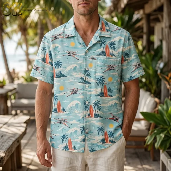 A man wears a light blue Star Wars Hawaiian shirt with white shorts on a wooden deck surrounded by tropical greenery.