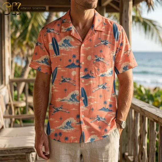 A man wears a coral pink spacecraft-print Hawaiian shirt with light-colored pants on a wooden deck overlooking the ocean.