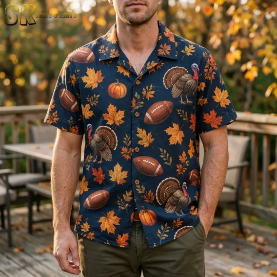 A man wears a navy Thanksgiving Hawaiian shirt with turkey and football illustrations on a wooden deck surrounded by fall foliage.