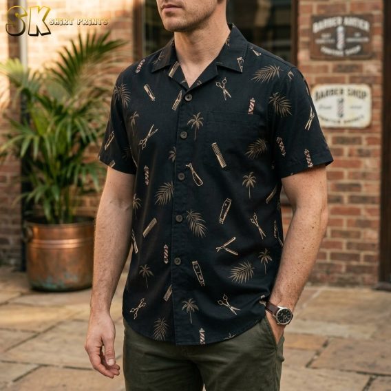 A man stands in front of a brick barbershop exterior wearing a black button-up shirt printed with scissors, hair clippers, combs, barber poles, and palm trees in beige and brown tones.