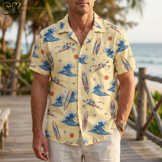 A man wears a pale yellow wave-and-spacecraft shirt with light shorts on a wooden boardwalk beside the ocean.