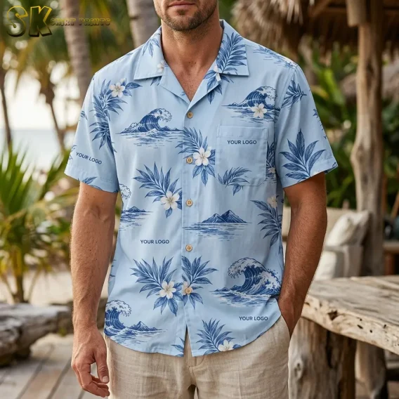 A man wearing a light blue custom logo Hawaiian shirt stands outdoors near a thatched structure, with the chest pocket and repeating wave and palm leaf print clearly visible from the front.