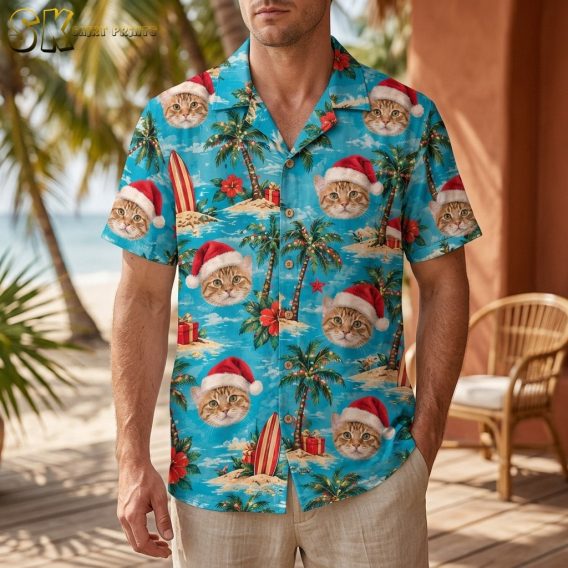 A man wearing a santa aloha shirt stands on a beach-side deck, the turquoise fabric covered in orange tabby cat faces under red Santa hats alongside palm trees and wrapped gifts.