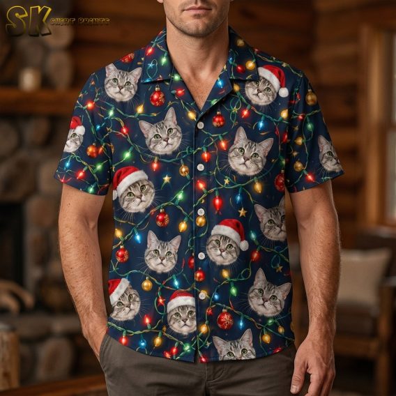 A man wearing a funny christmas hawaiian shirt printed with photorealistic cat faces in Santa hats and glowing multicolored string lights on a navy background, photographed from the chest down in a warm wood-paneled interior.