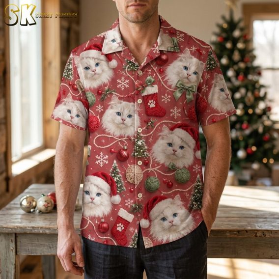 A man wearing a mens christmas hawaiian shirt stands in a warmly lit room with a decorated tree behind him, the fluffy white cats and red ornament motifs clearly visible across the chest and sleeves.