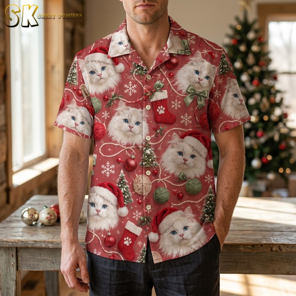A man wearing a mens christmas hawaiian shirt stands in a warmly lit room with a decorated tree behind him, the fluffy white cats and red ornament motifs clearly visible across the chest and sleeves.