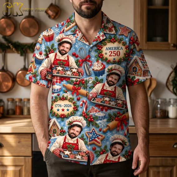 A bearded man wearing the light blue all-over printed shirt stands in a warm kitchen, with the 'AMERICA 250' wreath motif and repeated smiling chef figures clearly visible across the chest and sleeves.