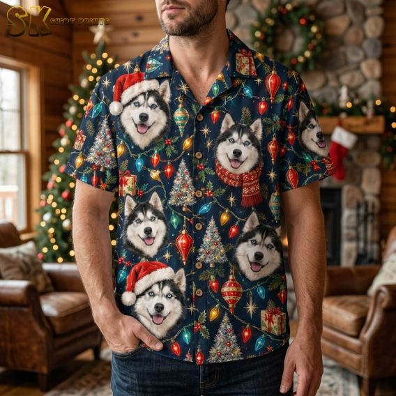 A man wearing the navy husky Christmas shirt stands in a cozy log-cabin living room with a decorated tree and fireplace behind him, the large husky faces and colorful string-light print clearly visible across the chest.
