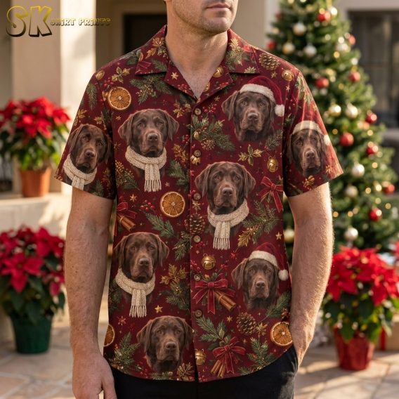 A man wearing a deep burgundy christmas hawaiian shirt printed with repeating chocolate Labrador portraits in Santa hats, standing in front of a decorated Christmas tree.