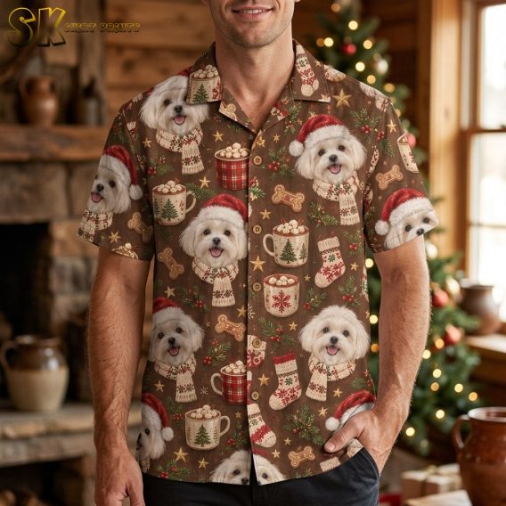 A smiling man wearing a mens christmas hawaiian shirt with white Maltese dog faces in Santa hats stands in front of a decorated Christmas tree inside a cozy log cabin.