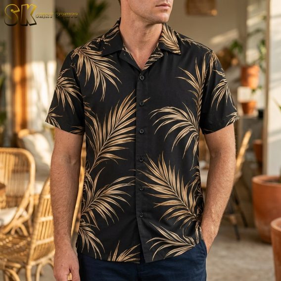 A man stands indoors wearing a black short-sleeve shirt covered in large beige palm fronds, with rattan furniture and potted plants visible in the background.