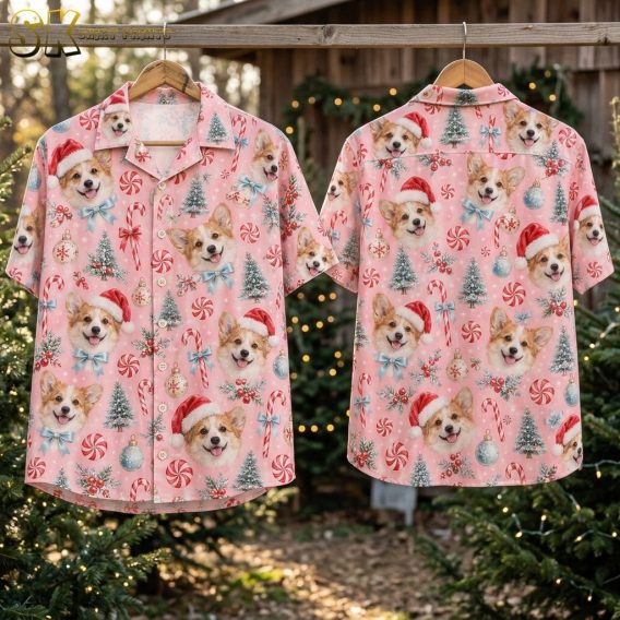 Front and back of the pink corgi Santa shirt hang on wooden hangers outdoors among evergreen trees strung with warm lights, showing the full repeat of dog faces, ornaments, and candy canes on both sides.