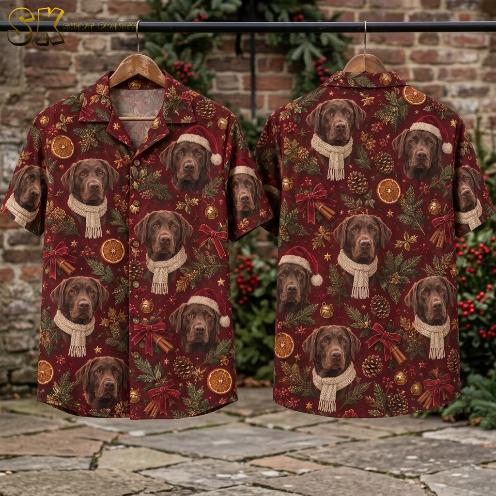 Front and back of the chocolate Lab Christmas shirt hanging on wooden hangers against a brick wall dressed with red berries and pine garland, showing the full repeat of Labrador portraits, pinecones, cinnamon sticks, and red bows across both panels.