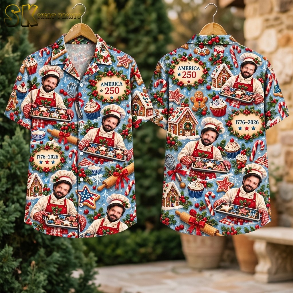 The shirt hangs on two wooden hangers outdoors against green foliage, showing the front with the smiling chef and 'AMERICA 250' wreath and the back with the gingerbread house centerpiece and '1776–2026' text.
