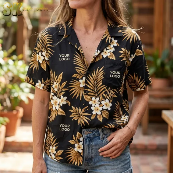A woman wearing a black custom logo Hawaiian shirt with large cream plumeria blooms and golden-brown palm fronds stands in a warm outdoor setting, with 'YOUR LOGO' text visible on the left chest pocket and lower front panel.